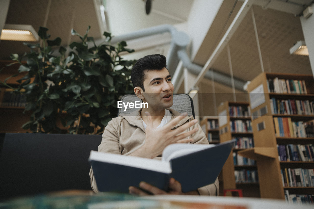 Male student gesturing while holding book in library