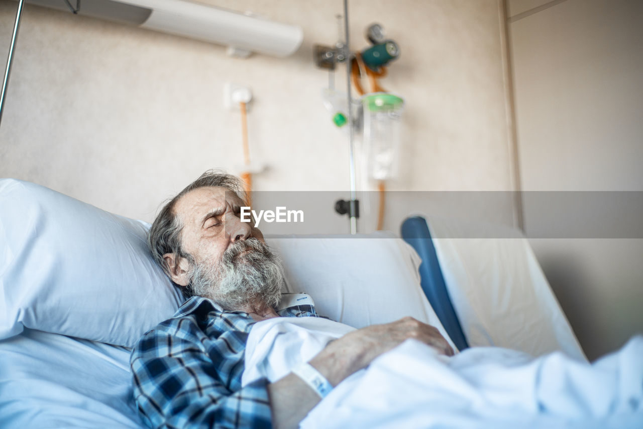 Calm aged man with beard lying under blanket on bed in hospital ward and sleeping