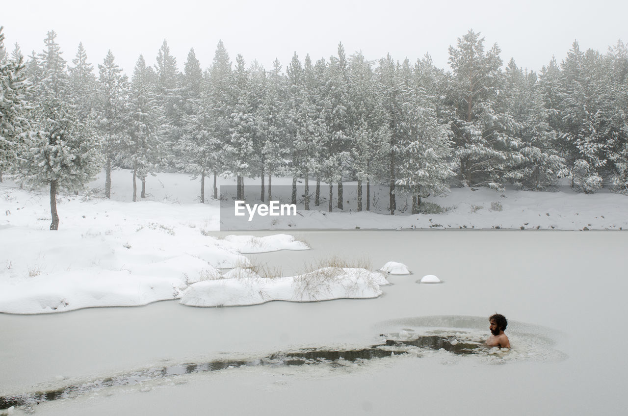 Snow covered land and trees during winter and a man swimming in a frozen lake