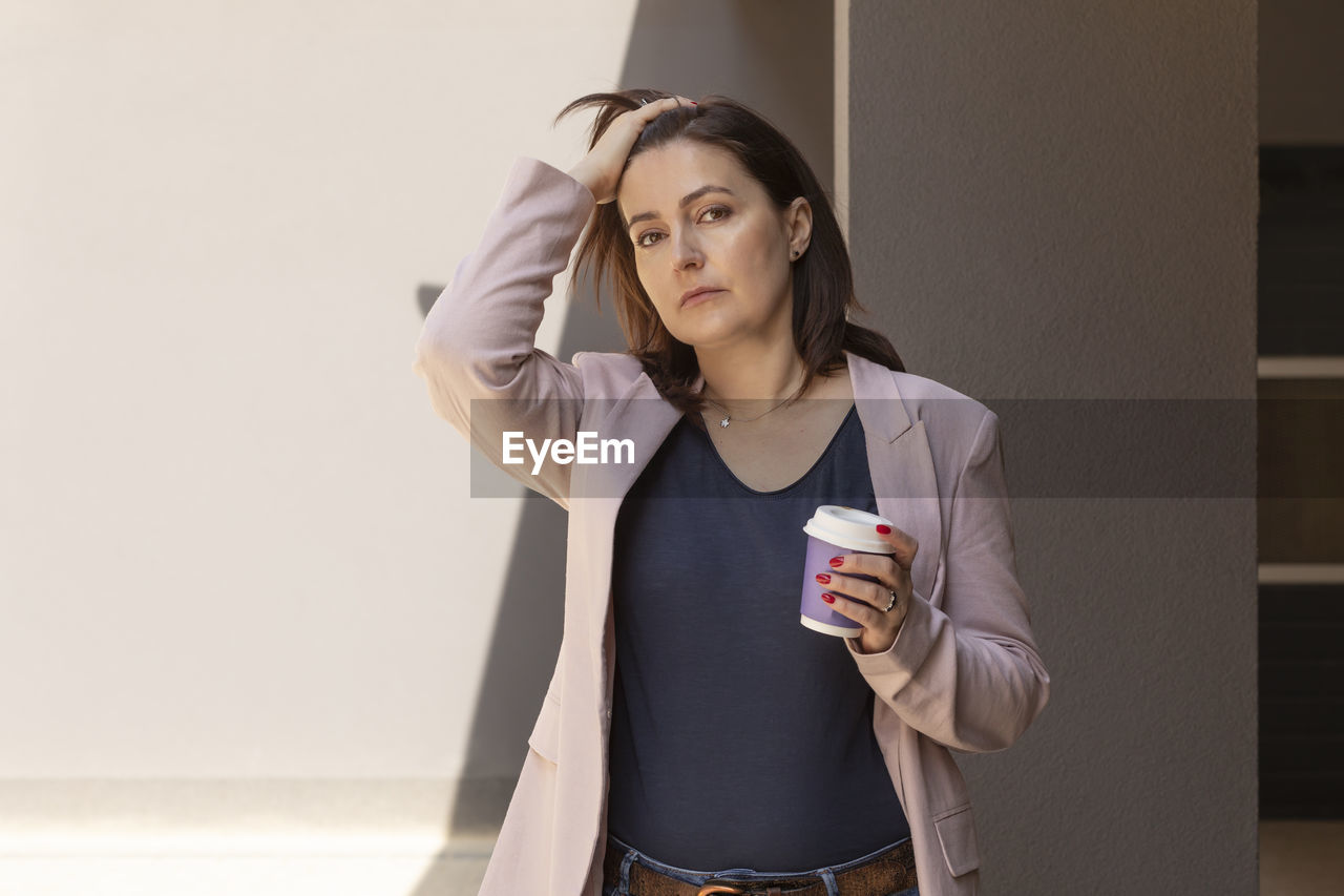 Puzzled sad mature 40 yo woman holds cup of coffee, looking at camera outside, tired, worried