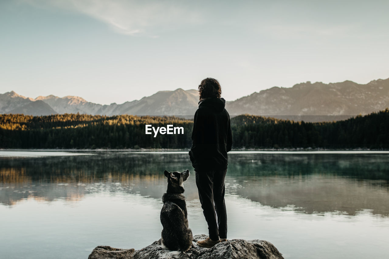 Rear view of man with dog looking at lake against mountain range