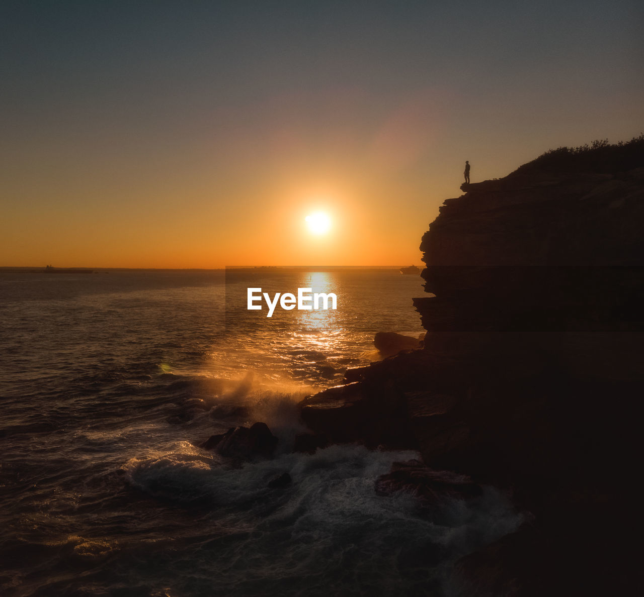 Drone aerial of silhouette of person standing at coastal cliff edge during golden hour sunset