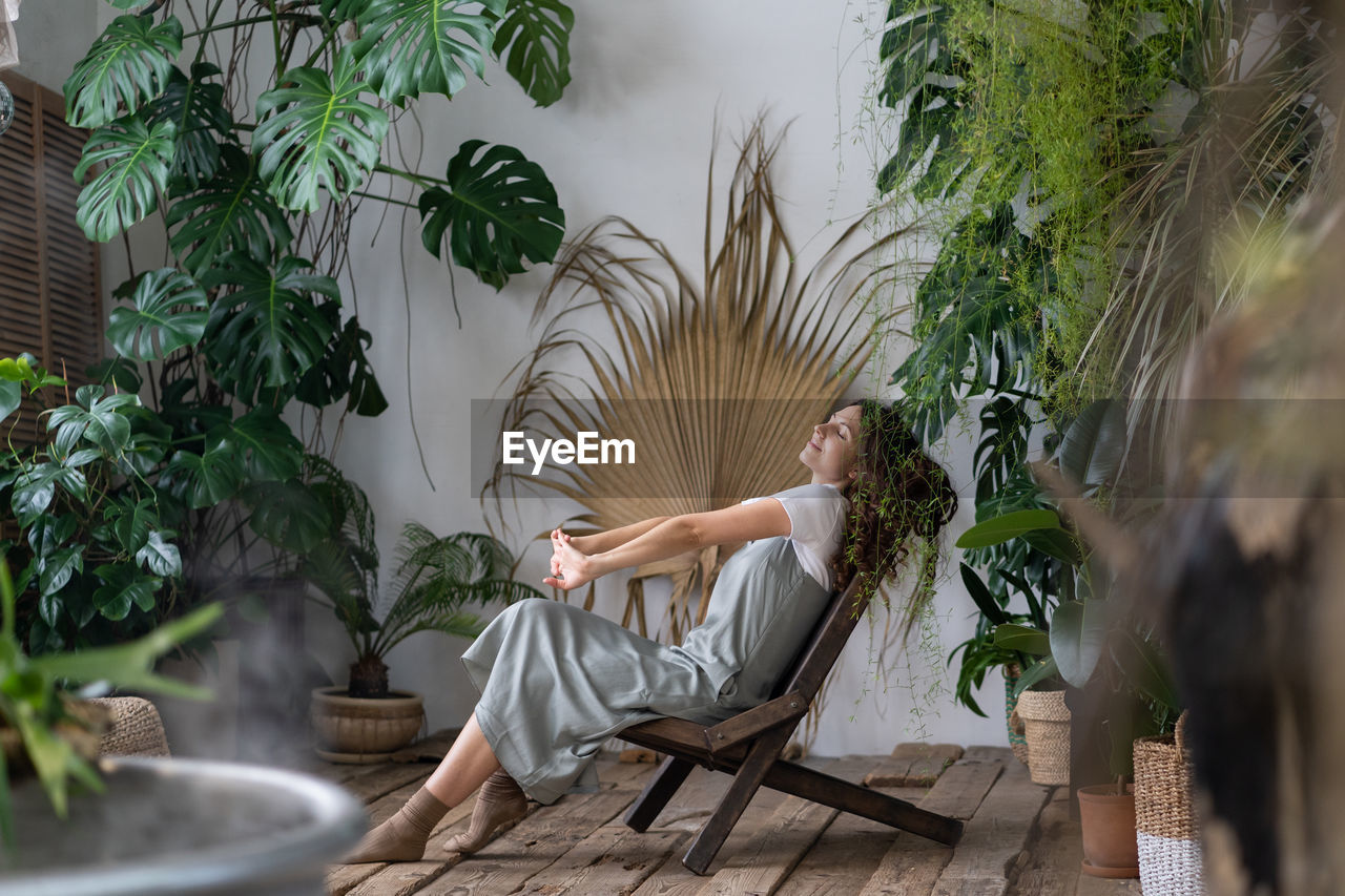 Dreamy woman relaxing in tropical resort-style garden at home, female gardener resting in greenhouse