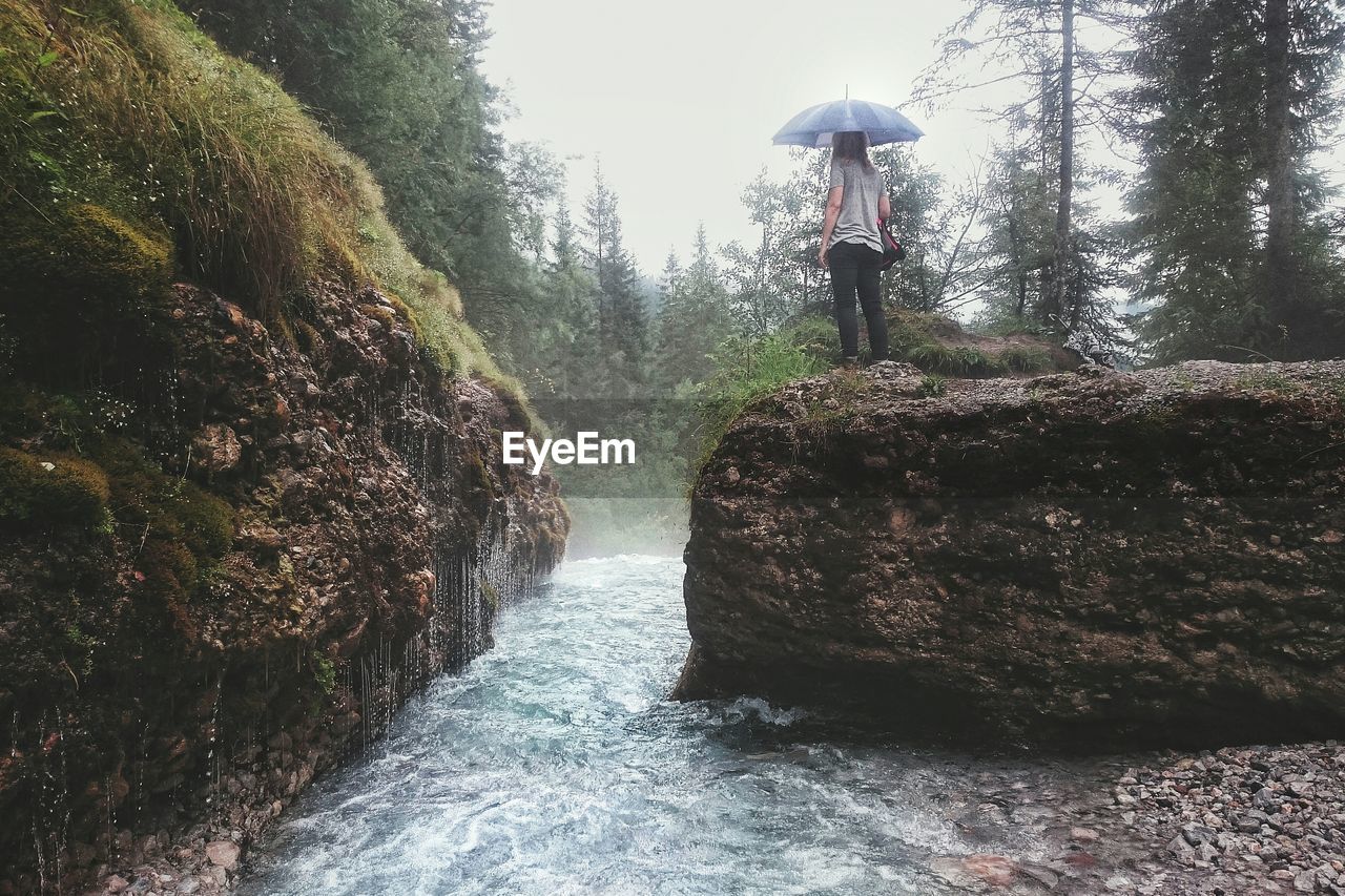 Rear view of woman standing with umbrella on rock formation by river