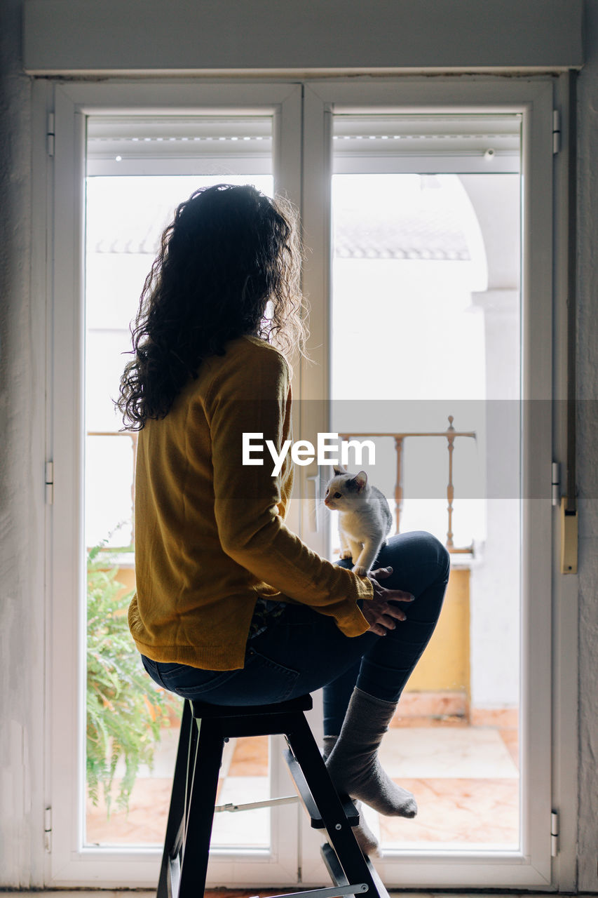 Full body side view of tranquil female in casual clothes sitting on stool near window and holding little kitten on knees while chilling at home