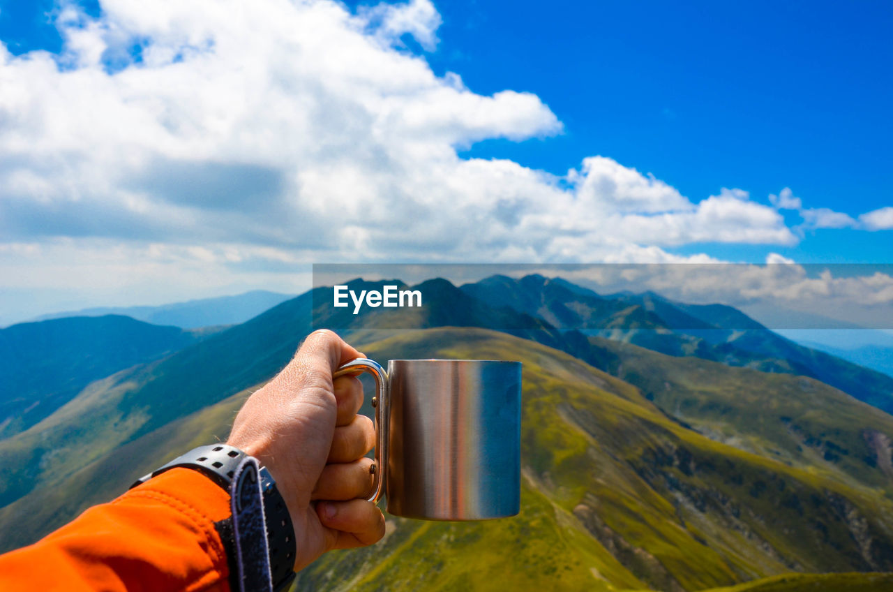 Cropped image of hand holding mug towards mountain range against sky