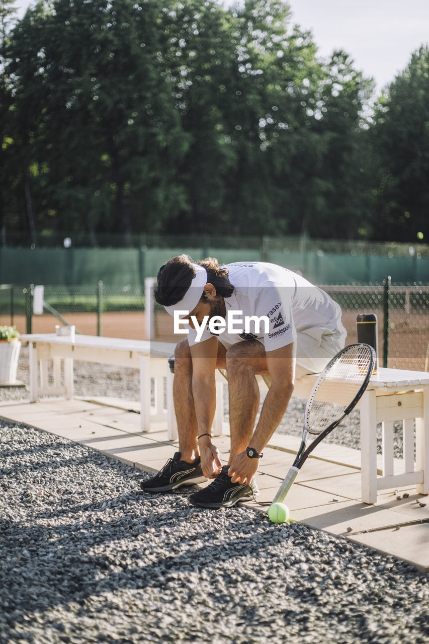 Man tying shoe lace while sitting on bench at tennis court