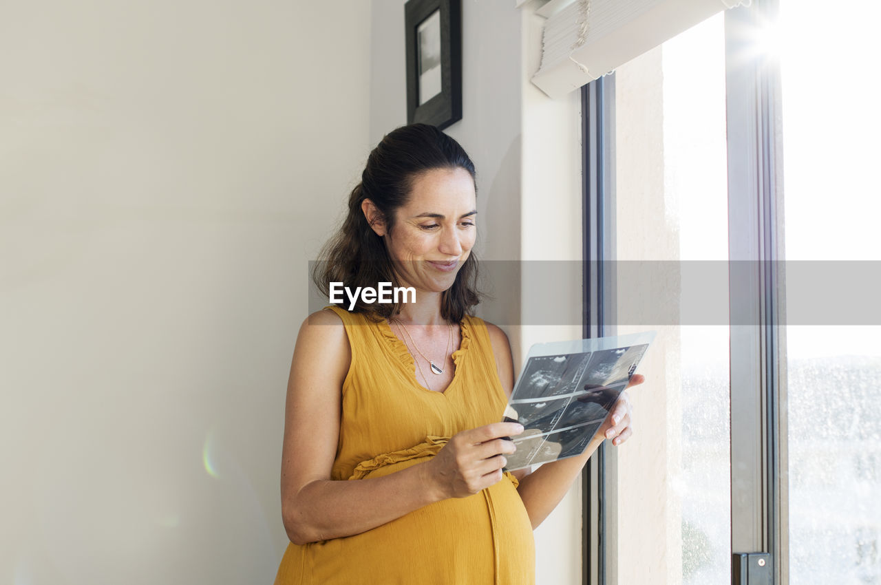 Smiling pregnant woman looking at x-ray while standing by window