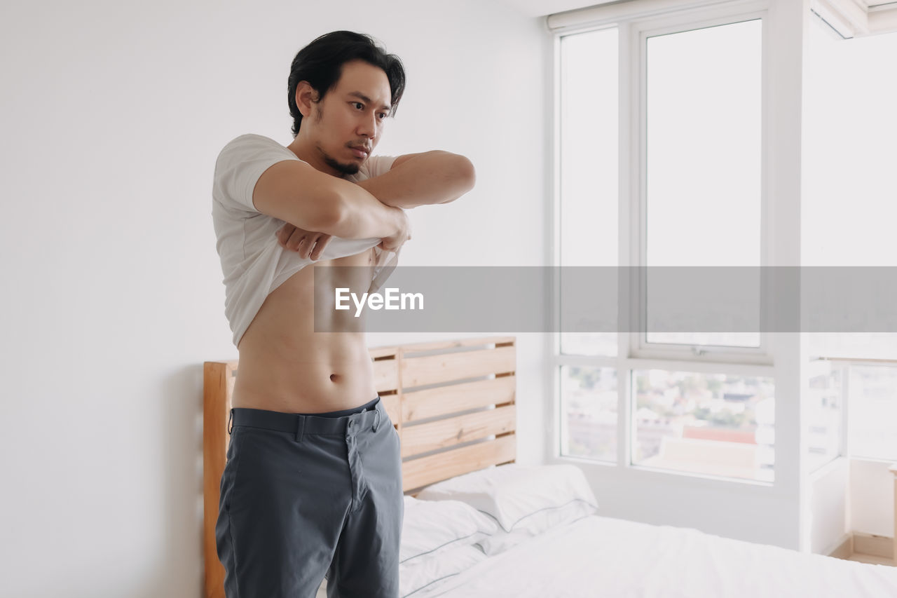 Young man looking away while standing on bed at home