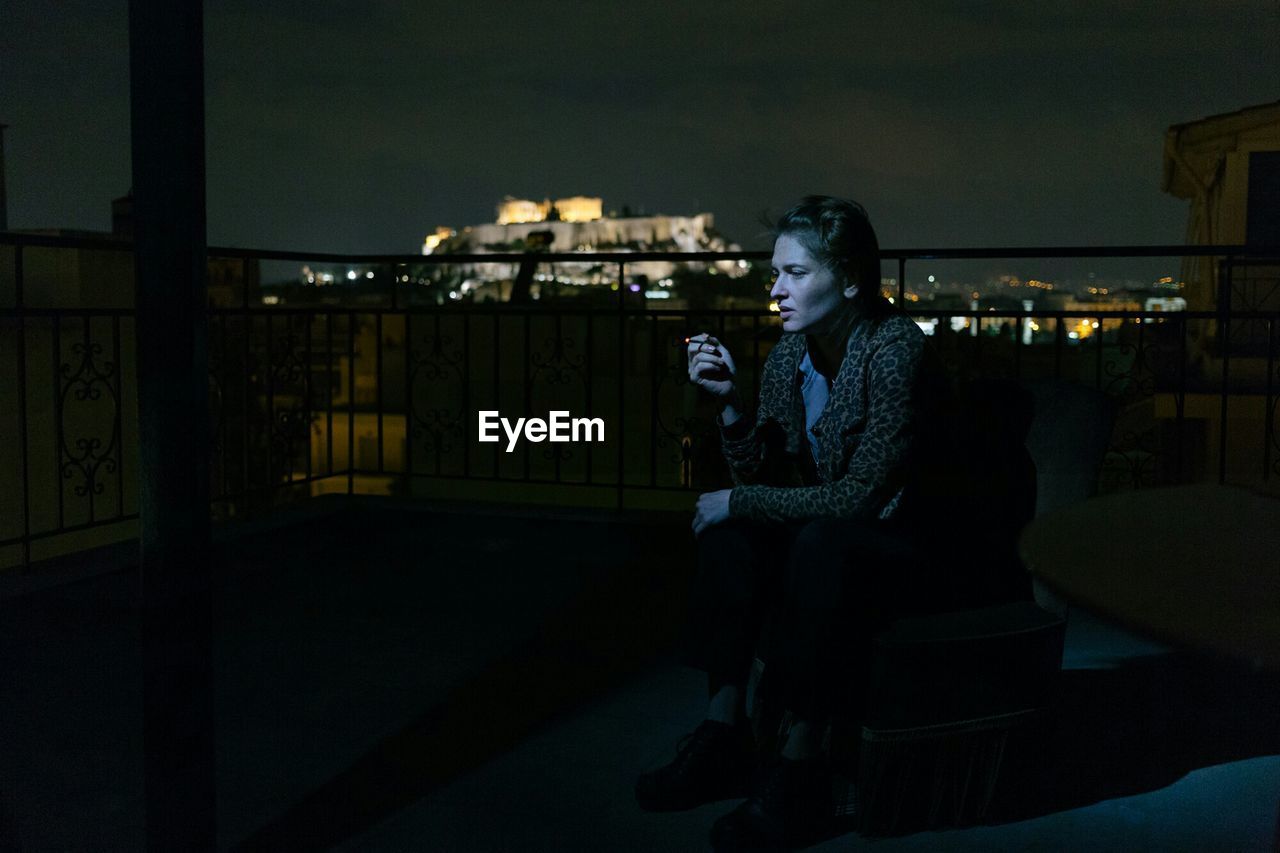 WOMAN SITTING ON ILLUMINATED BUILDING AGAINST SKY