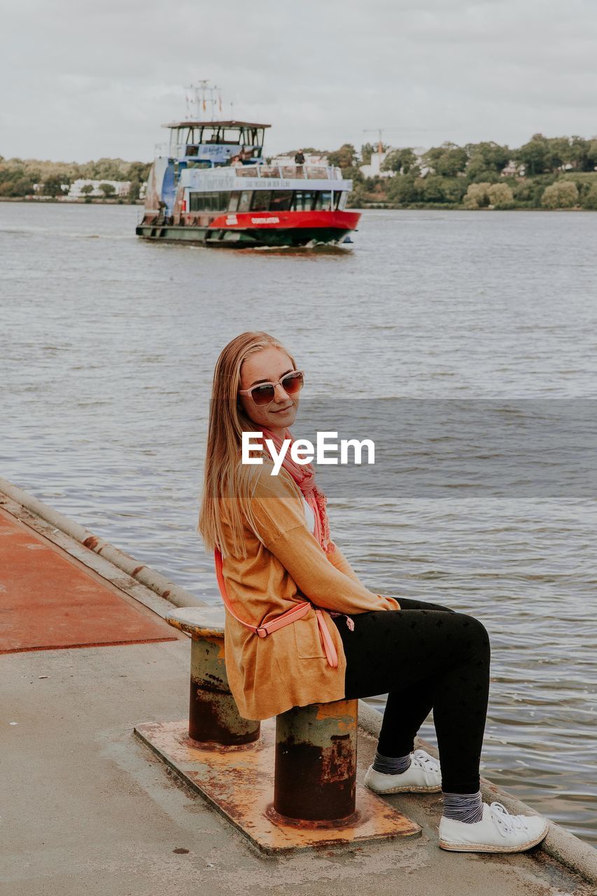 Portrait of young woman sitting on bollard against ferry in river