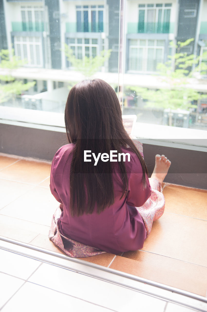 Rear view of girl sitting on tiled floor by window at home