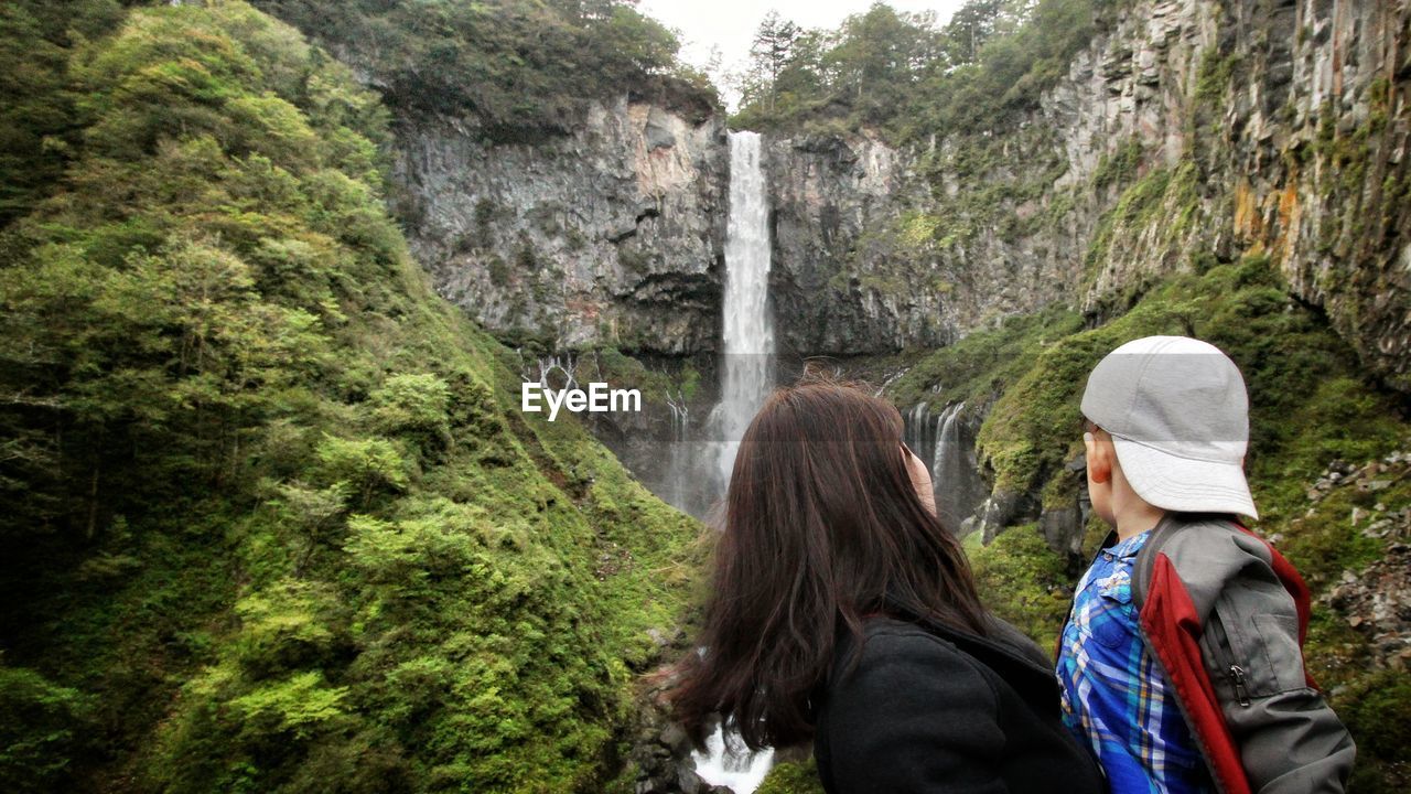 REAR VIEW OF WOMAN LOOKING AT WATERFALL ON ROCK