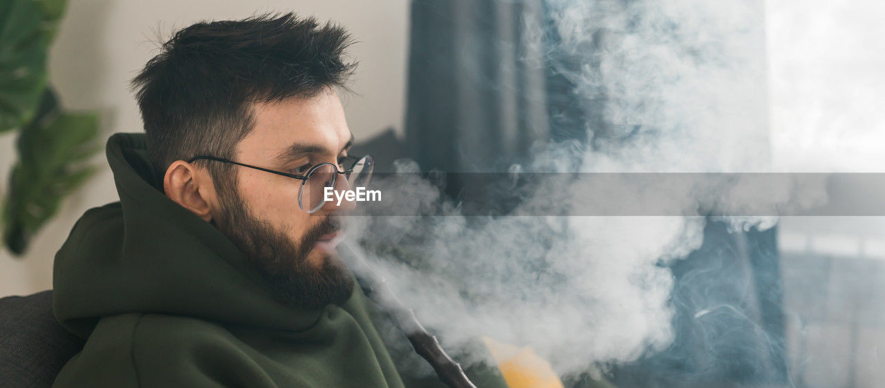 one person, adult, smoke, men, beard, glasses, facial hair, eyeglasses, portrait, headshot, person, human face, young adult, activity, smoking, lifestyles, nature, communication, looking, black hair, blue, sky, casual clothing, side view, outdoors, clothing