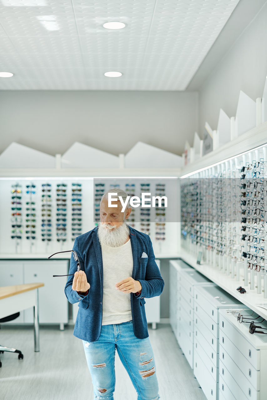 Serious elderly bearded male in trendy outfit standing with modern stylish glasses in optical shop