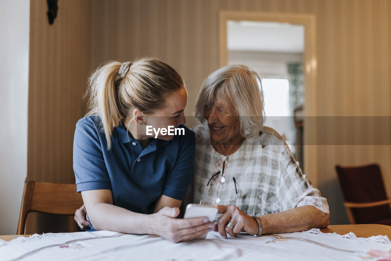 Smiling female care assistant sharing smart phone with senior woman while sitting at dining table in home