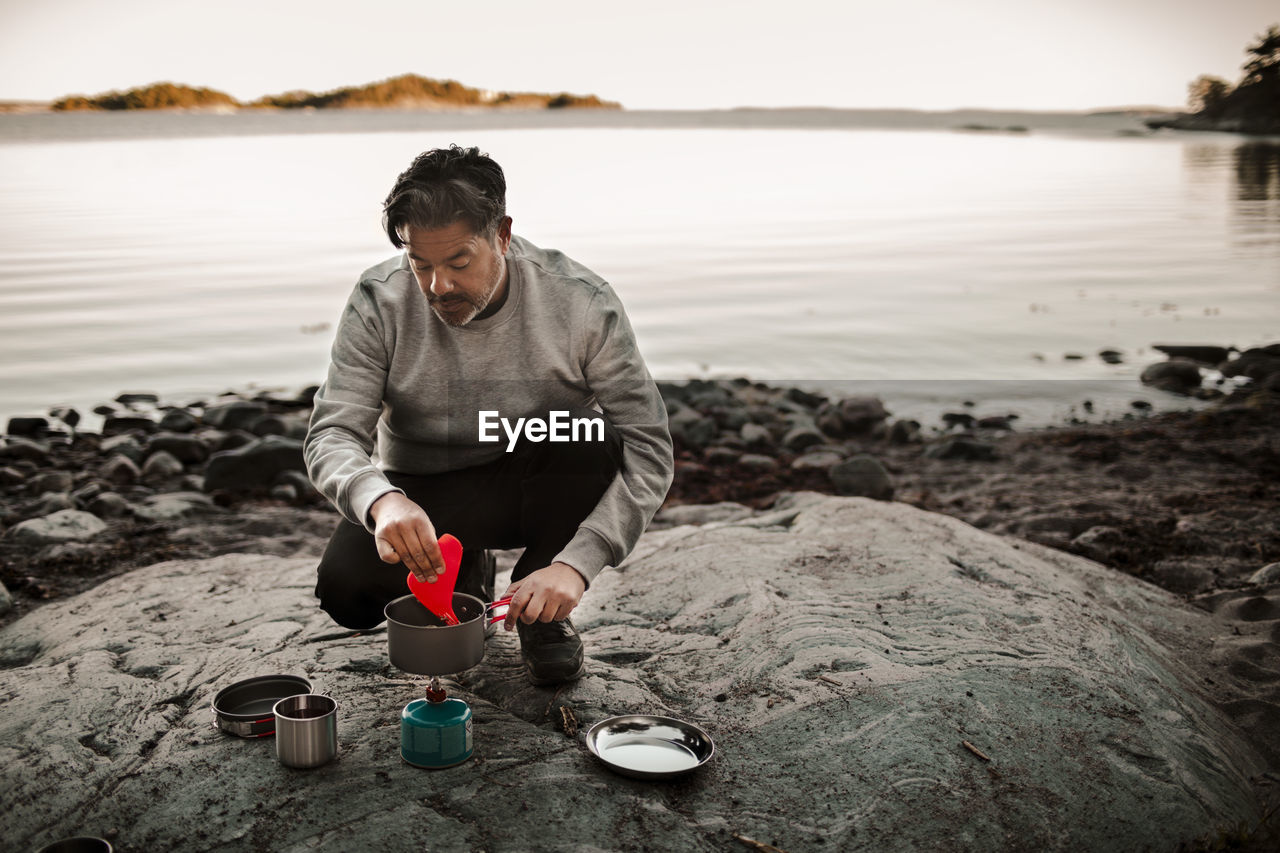 Full length of mature man cooking food on camping stove at lakeshore