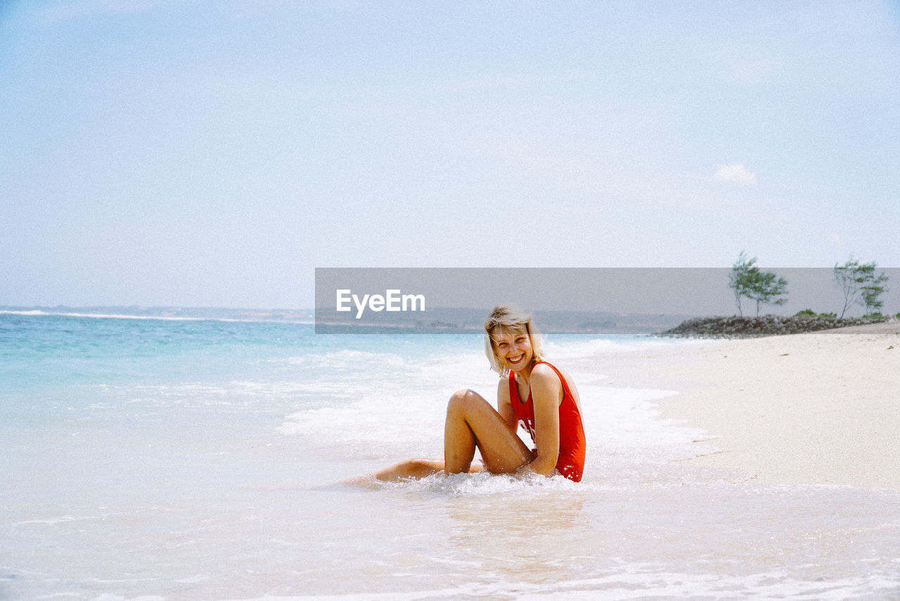 Portrait of smiling young woman sitting at beach against sky