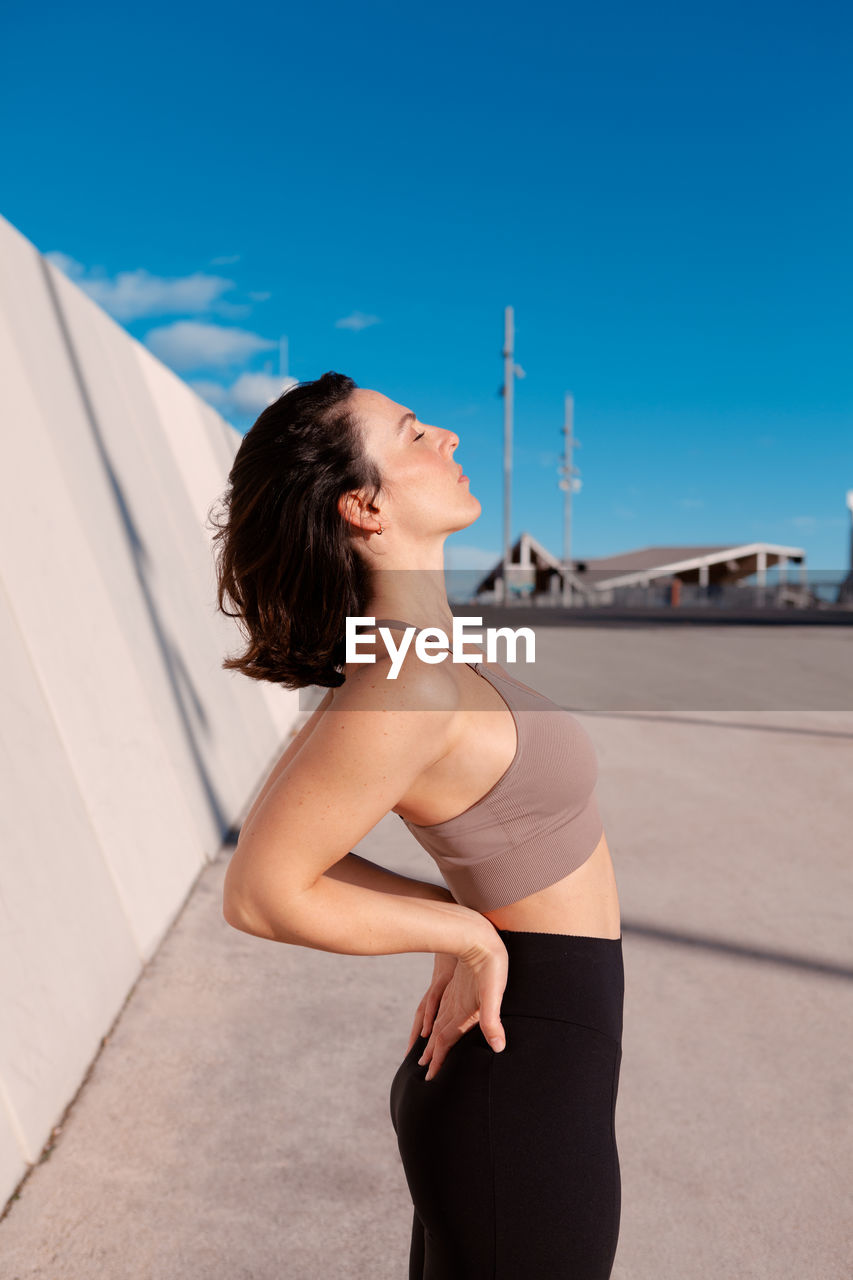 Side view of fit female in sportswear standing back bending while resting with eyes closed on street against blurred background during training on sunny summer day