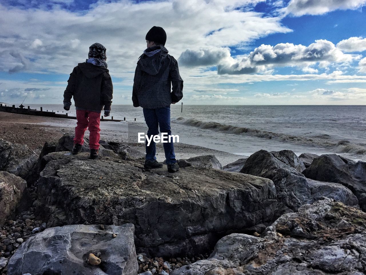 Rear view of brothers standing on rock at beach against cloudy sky during sunny day