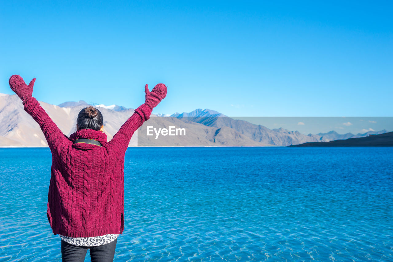 Woman standing by lake with arms raised against clear blue sky