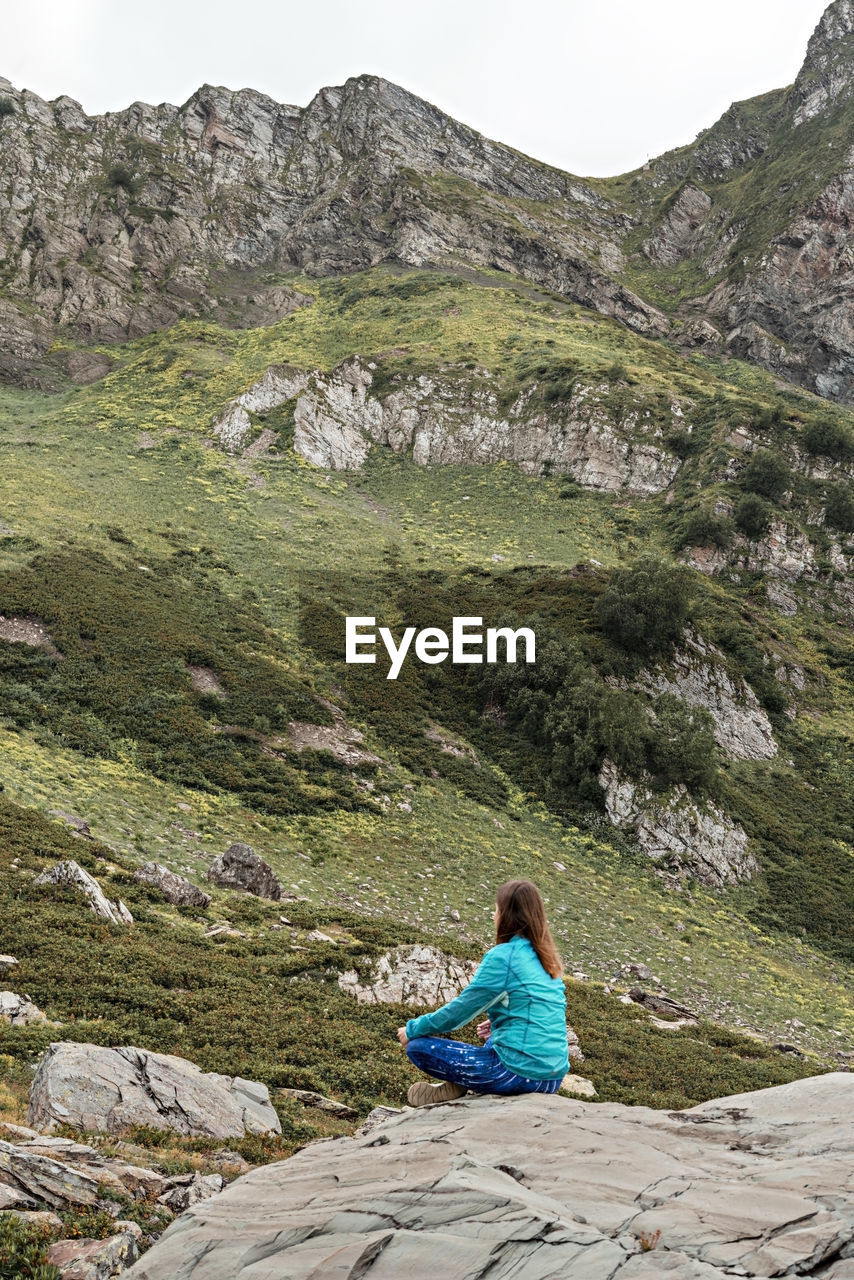 Rear view of meditating young woman in blue sitting on stone in mountain valley, consciousness