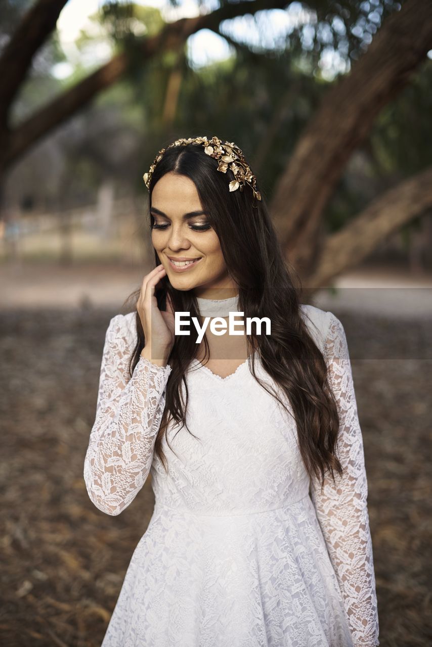 Charming tender young brunette in stylish white lace wedding dress and bridal wreath touching cheek and smiling while standing near tree in park