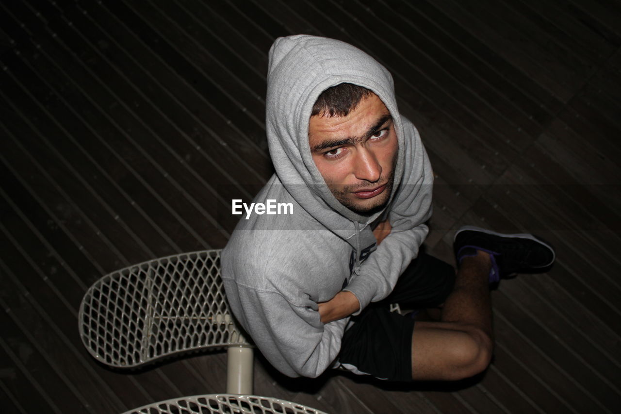 High angle portrait of hooded young man sitting on hardwood floor at home