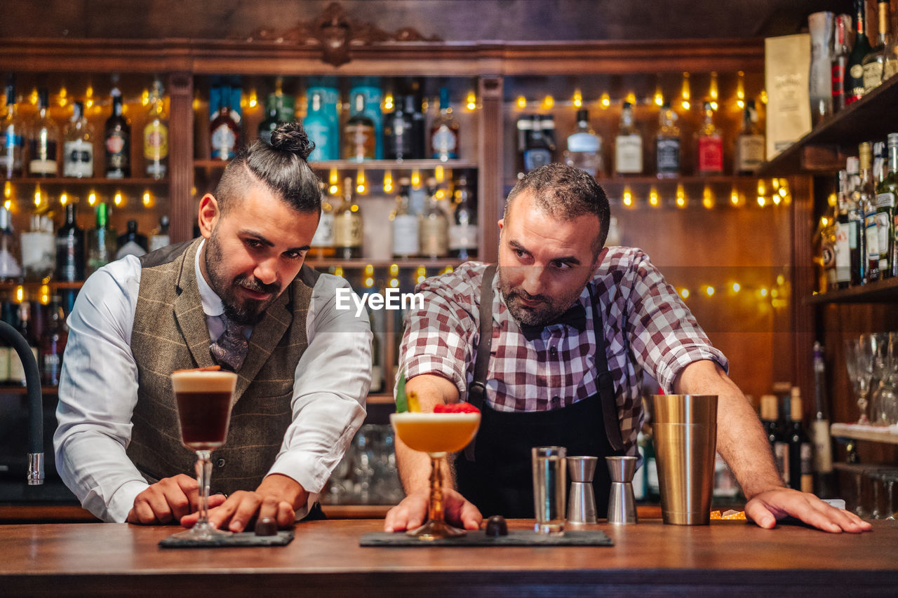 Bearded bartenders smiling while holding glasses of fresh cold sour cocktails at counter in bar