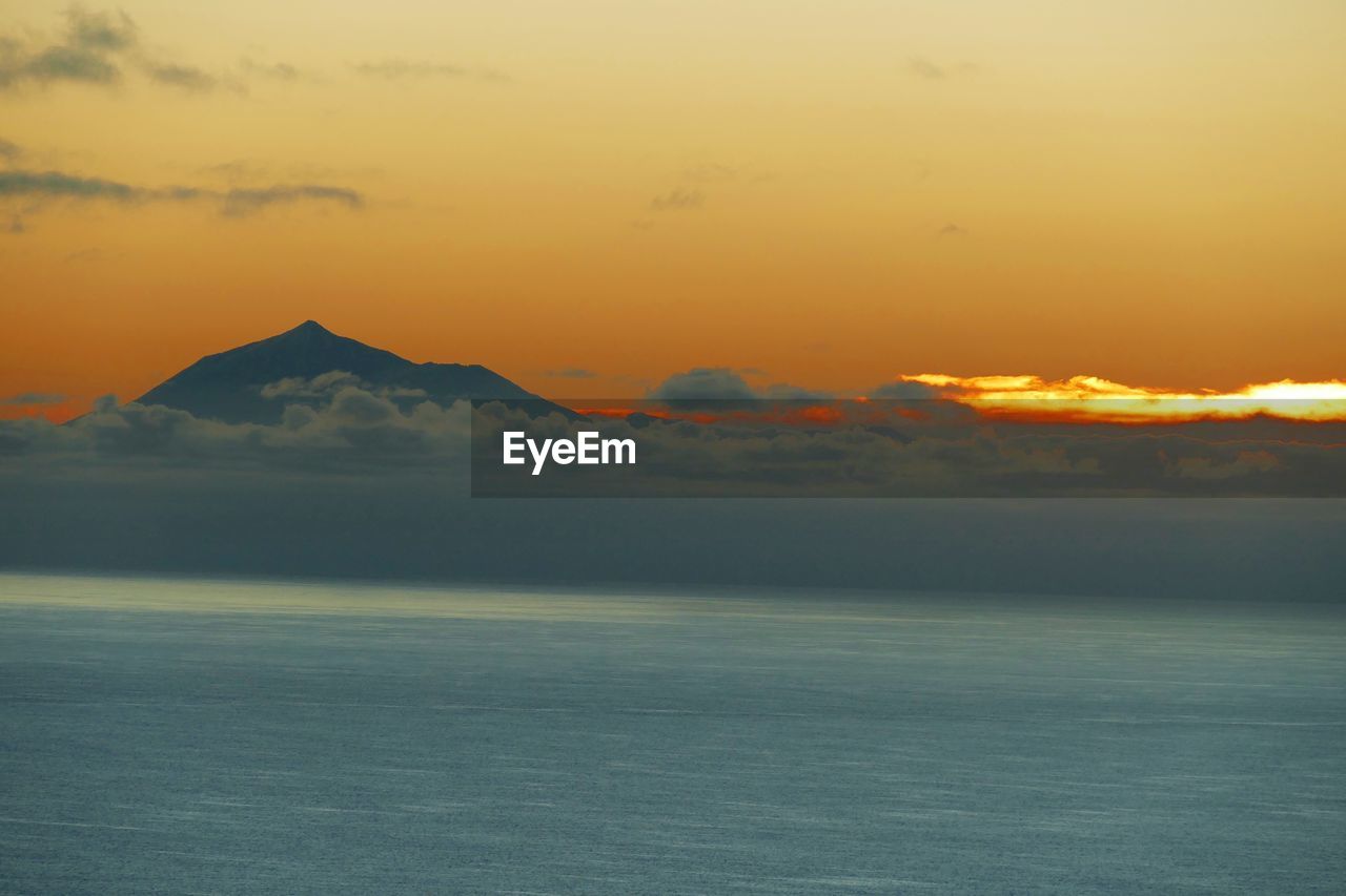SCENIC VIEW OF SEA AND SILHOUETTE MOUNTAINS AGAINST ORANGE SKY