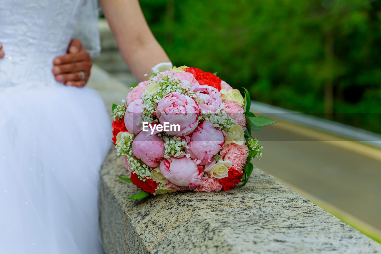 Cropped hands of groom holding bride waist by bouquet on retaining wall