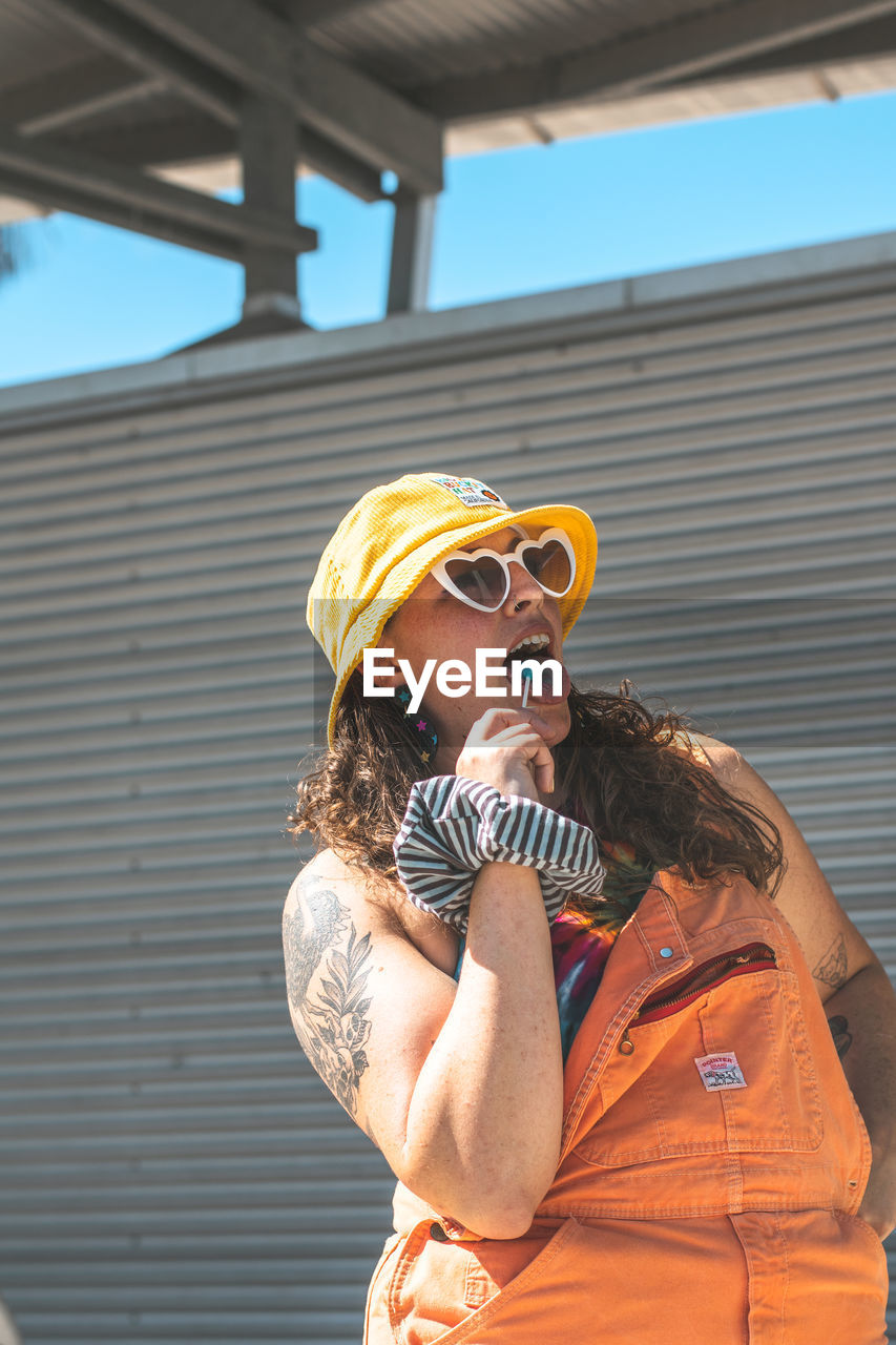 Portrait of young woman wearing sunglasses while standing against sky holding a lollipop