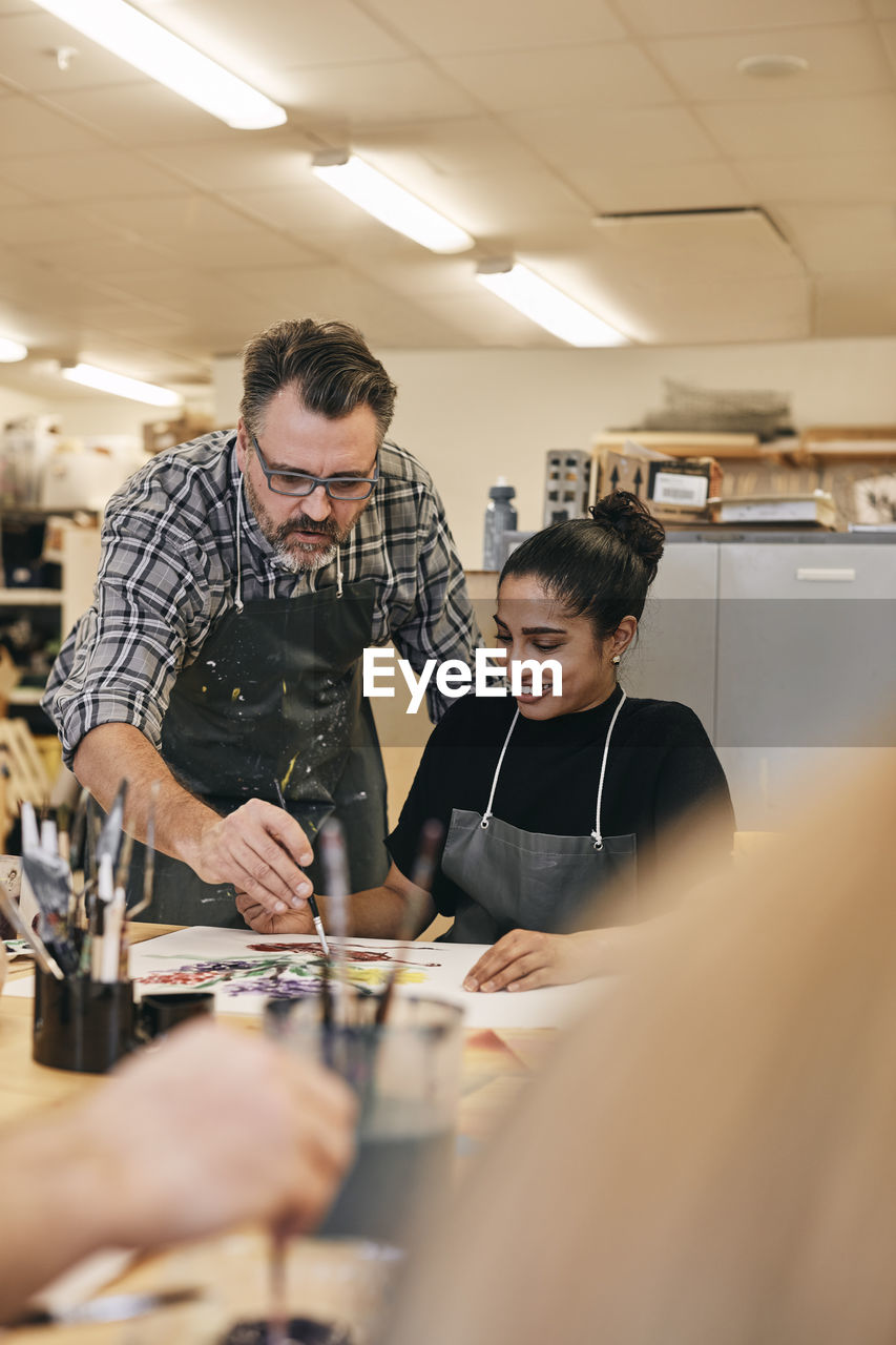 Instructor with paintbrush teaching student sitting at table in art class