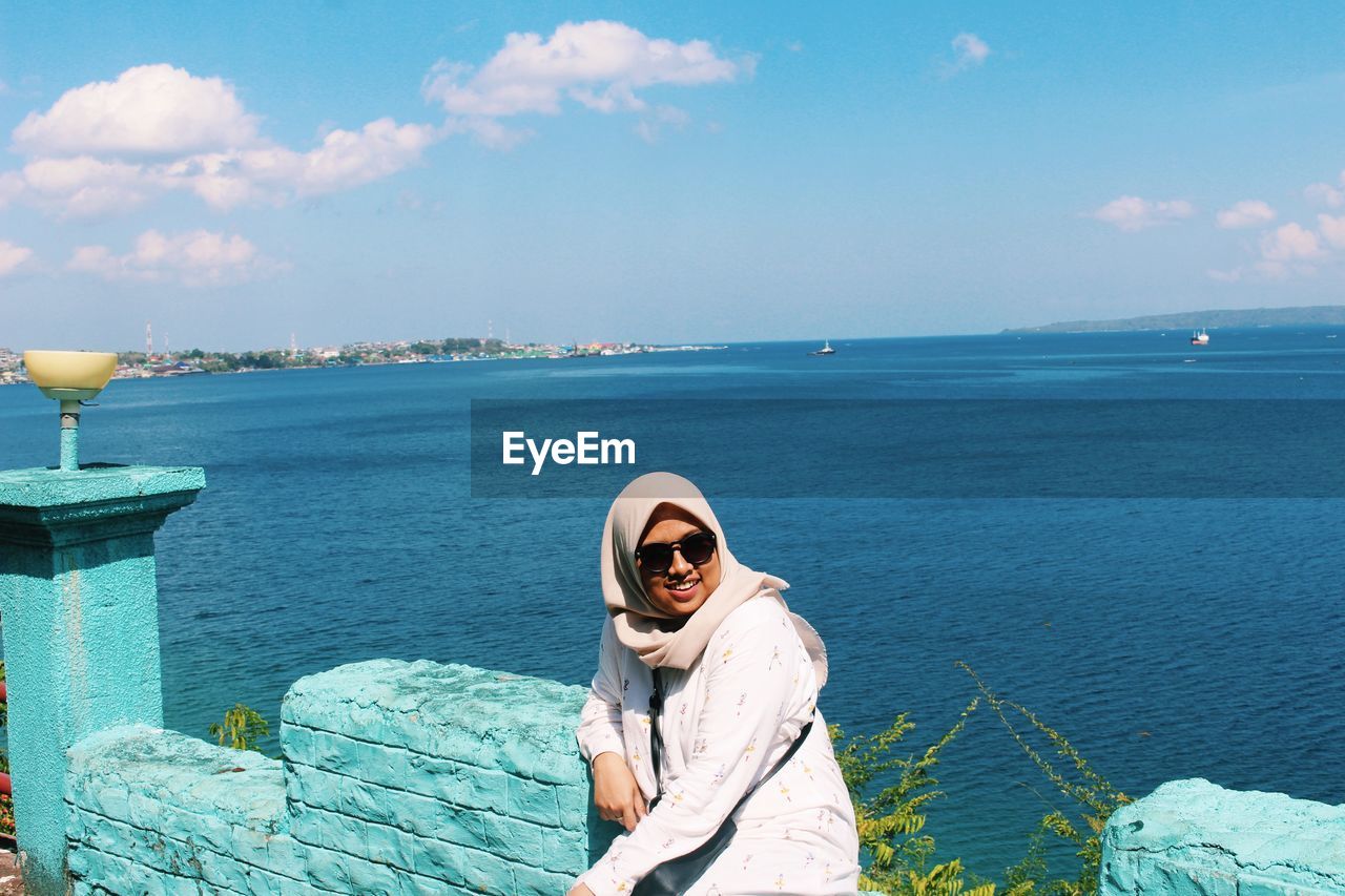 High angle view of young woman in hijab wearing sunglasses while sitting against sea during sunny day