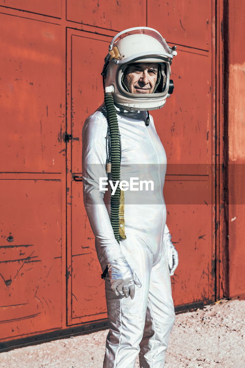 Man in spacesuit standing near red wall of industrial facility on sunny day