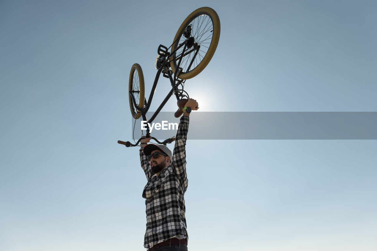 Low angle view of man with arms raised carrying bicycle while standing against clear blue sky during sunny day