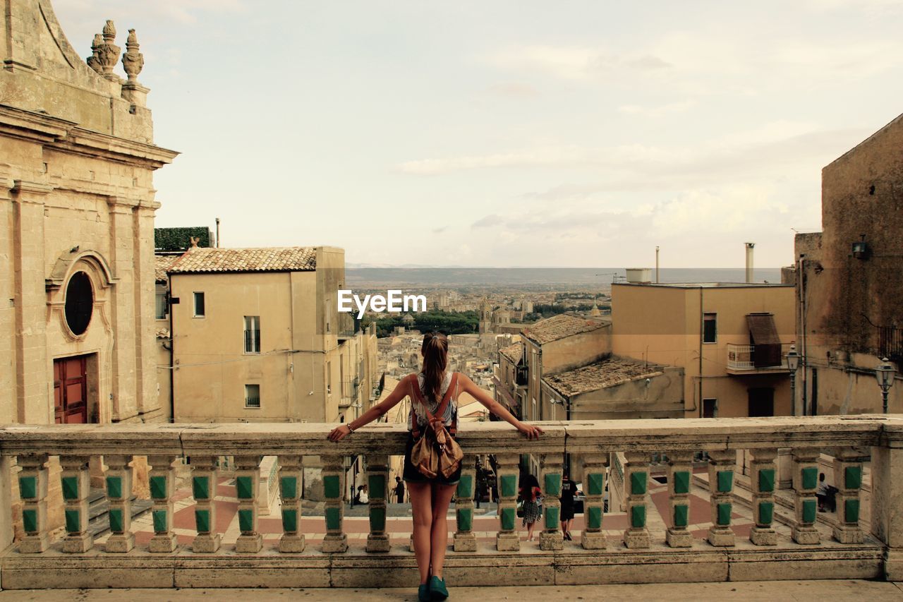 Rear view of woman standing by railing looking at city against sky