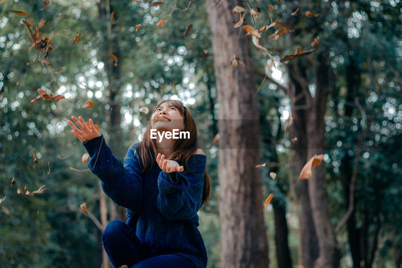 YOUNG WOMAN LOOKING AWAY WHILE SITTING ON TREE IN FOREST