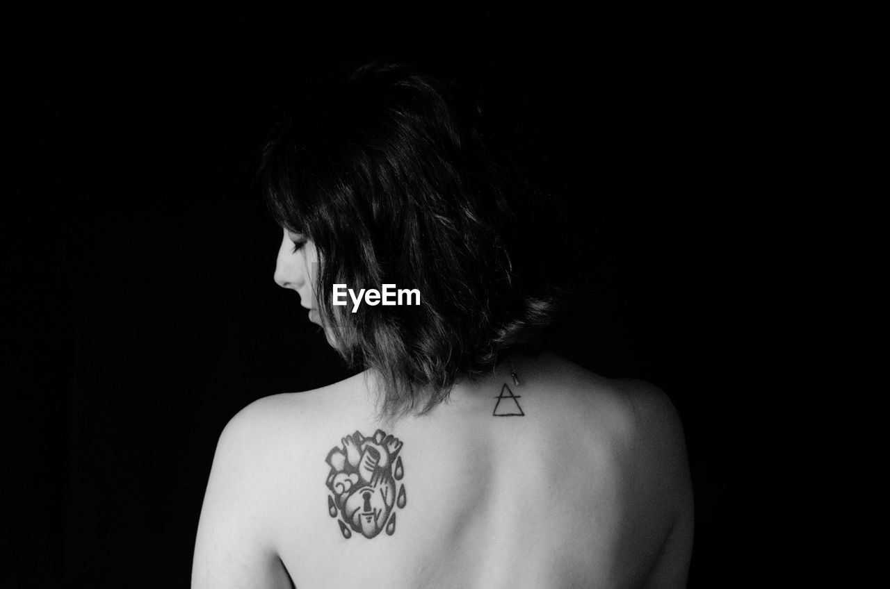 Rear view of shirtless woman with tattoos on back against black background