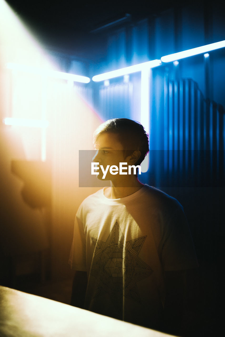 YOUNG MAN STANDING AGAINST ILLUMINATED LIGHT