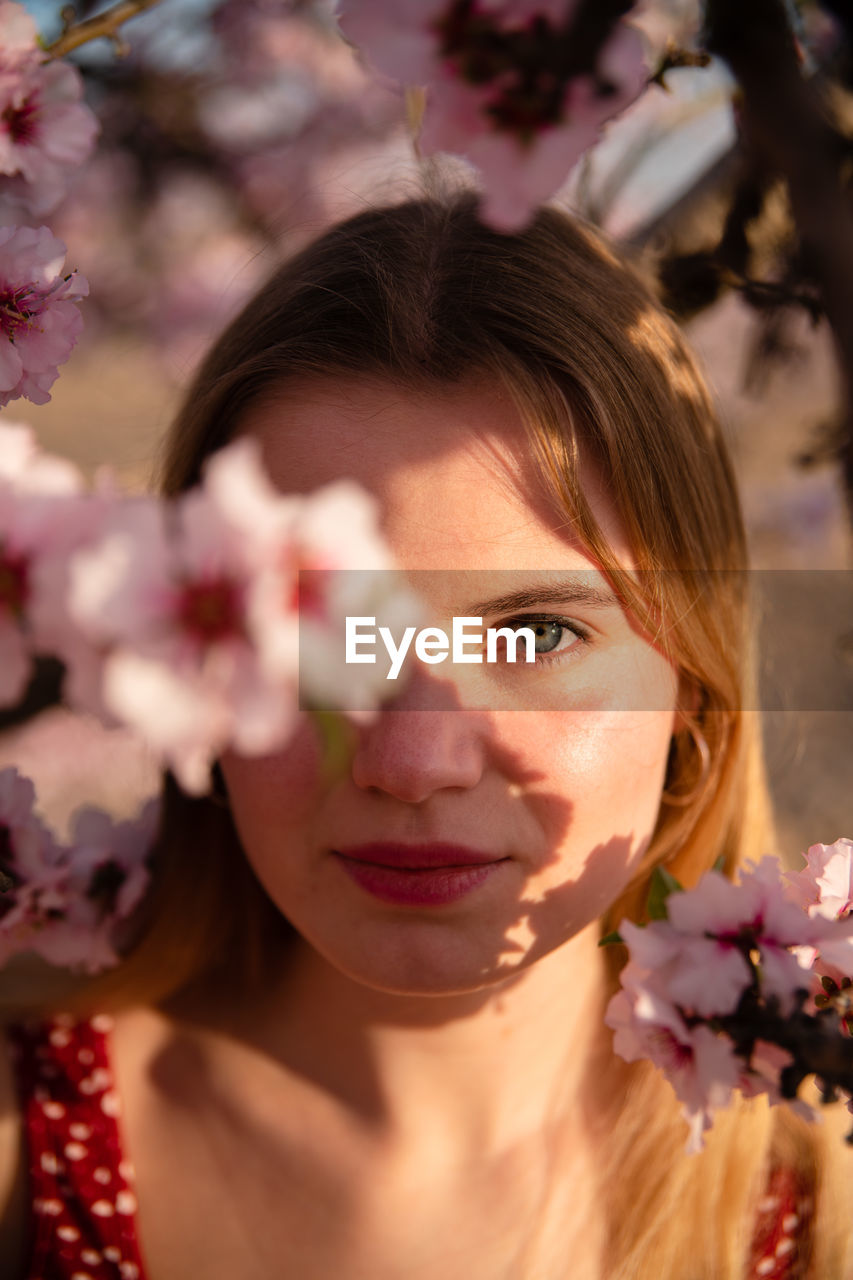 Blond woman with long hair posing under a flowering almond tree and looking at camera