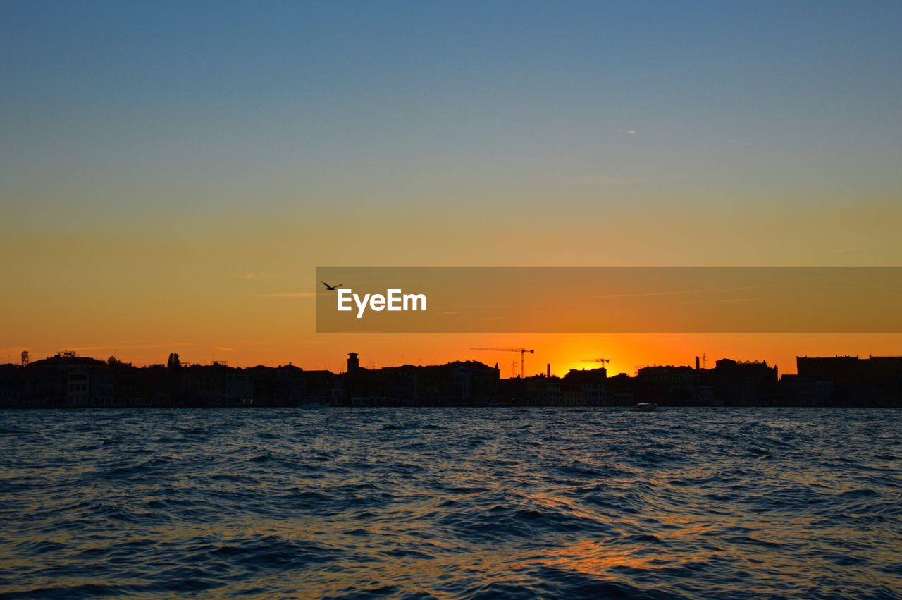 Sea by silhouette city against clear sky during sunset