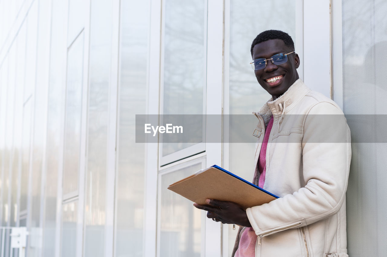 Stylish cheerful african american male entrepreneur reading documents on clipboard while standing on background of contemporary office building looking away