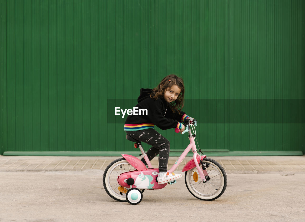 Girl riding bicycle on footpath against green wall