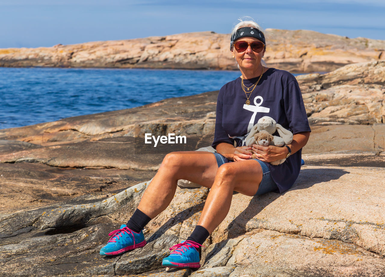 Full length portrait of senior woman with toys sitting on rock at beach during sunny day