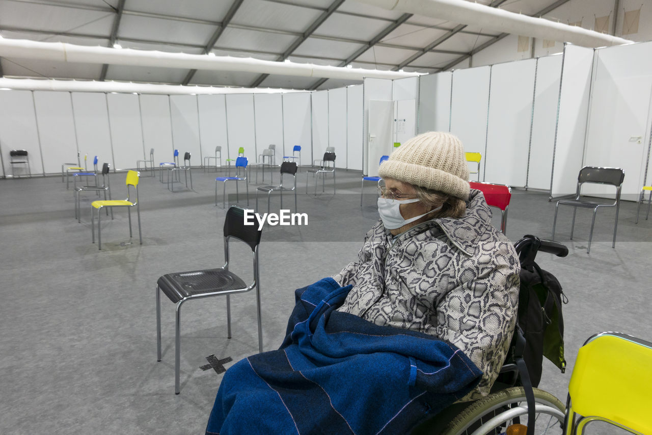 Senior women wearing protective face mask and warm clothing sitting on wheelchair at vaccination center