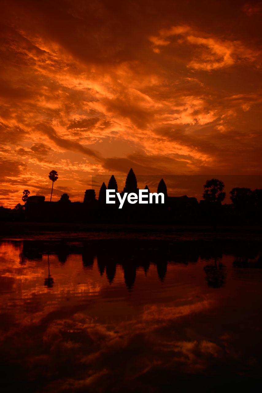 Reflection of silhouette built structure and trees in lake against orange sky during sunset