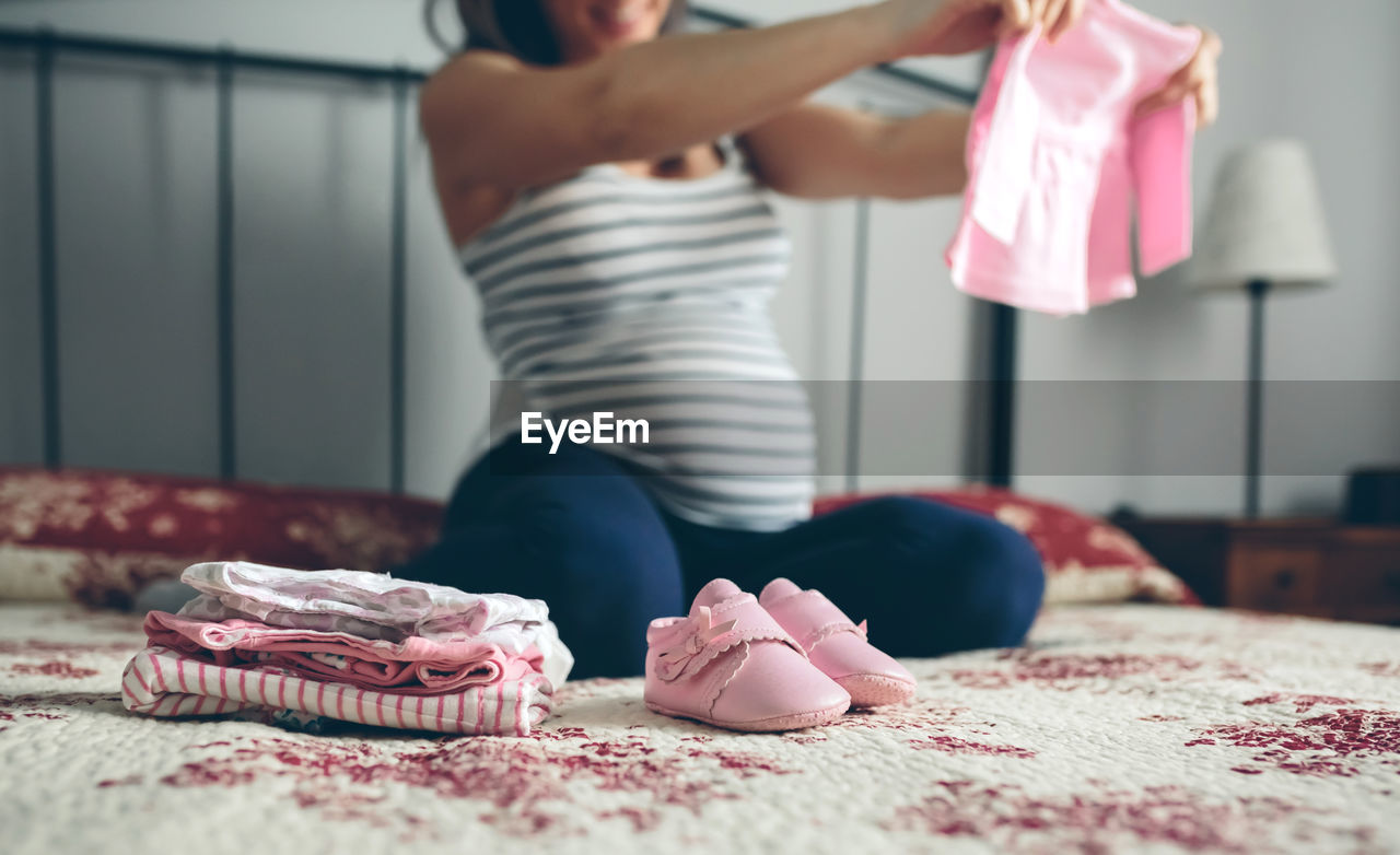 Pregnant woman holding baby clothing while kneeling on bed at home