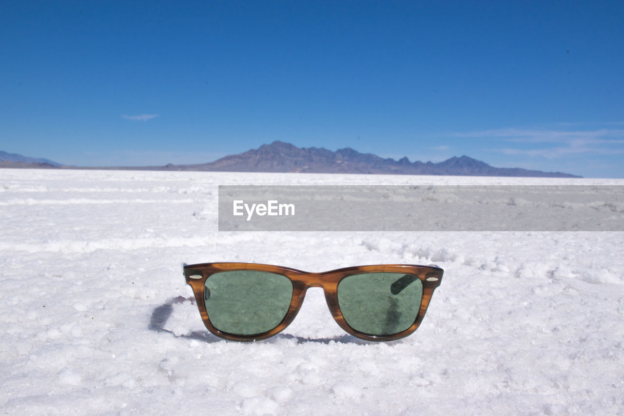 SUNGLASSES ON SAND AT BEACH AGAINST CLEAR BLUE SKY