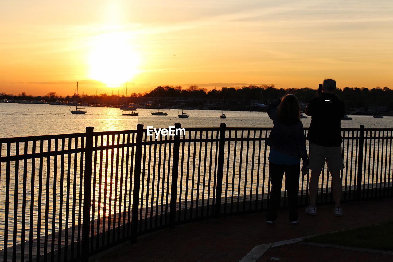 REAR VIEW OF PEOPLE STANDING BY RAILING AGAINST SUNSET SKY